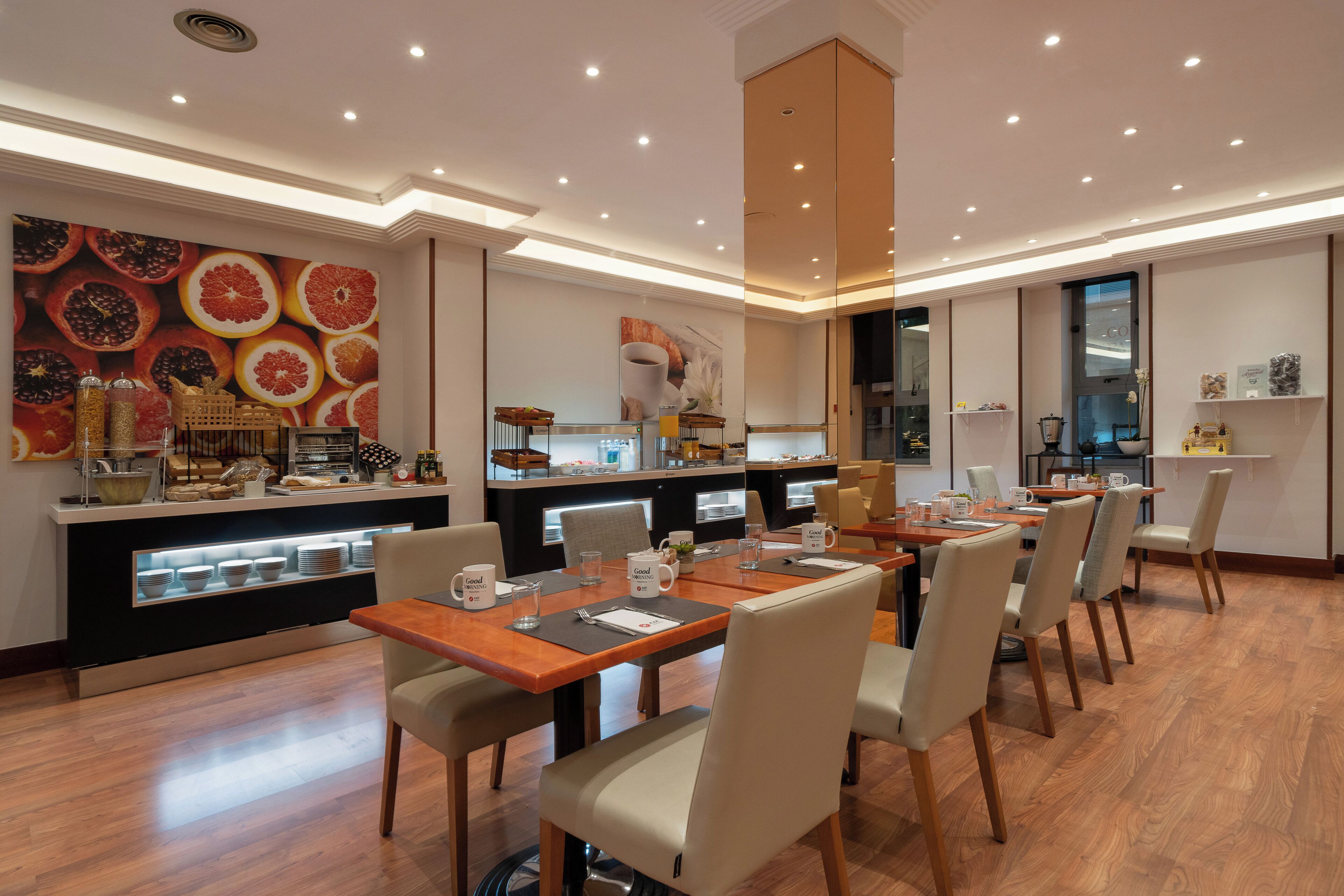 A modern dining area with a long wooden table, cushioned chairs, and a kitchen in the background.
