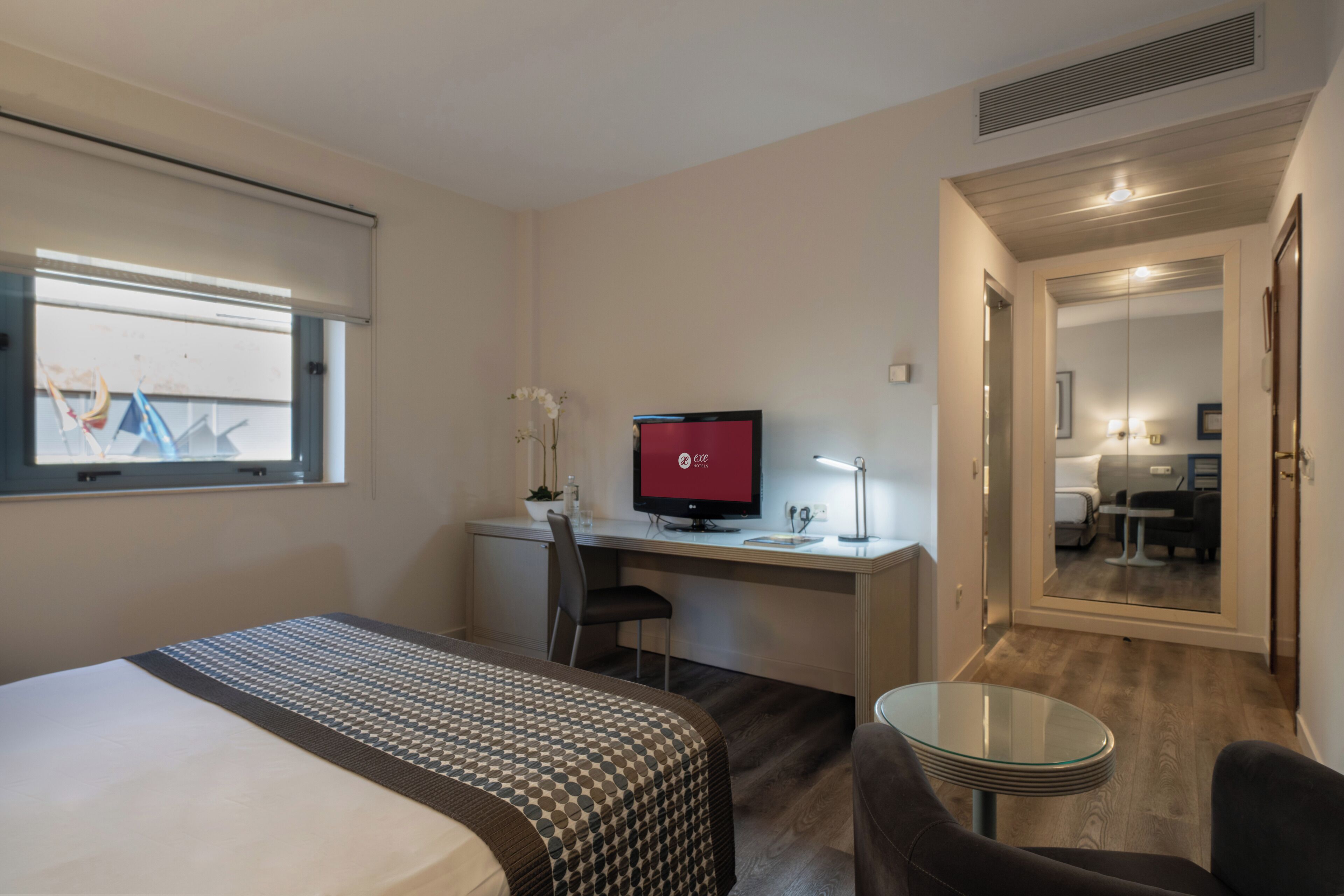 A hotel room with a bed, a desk with a computer, a chair, a small table, and a mirror.