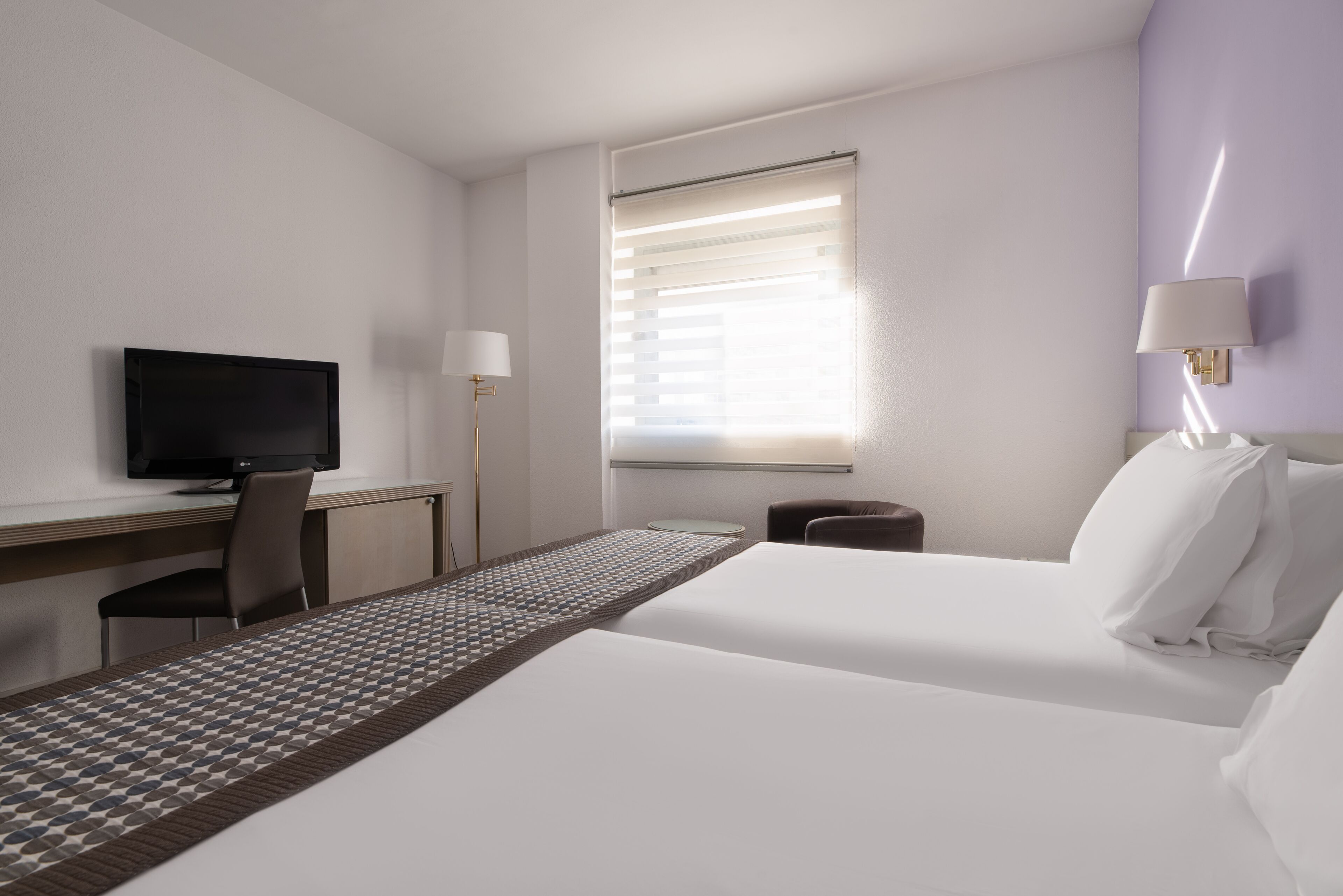 A hotel room with a bed, a desk, a television, a lamp, and a window with blinds.