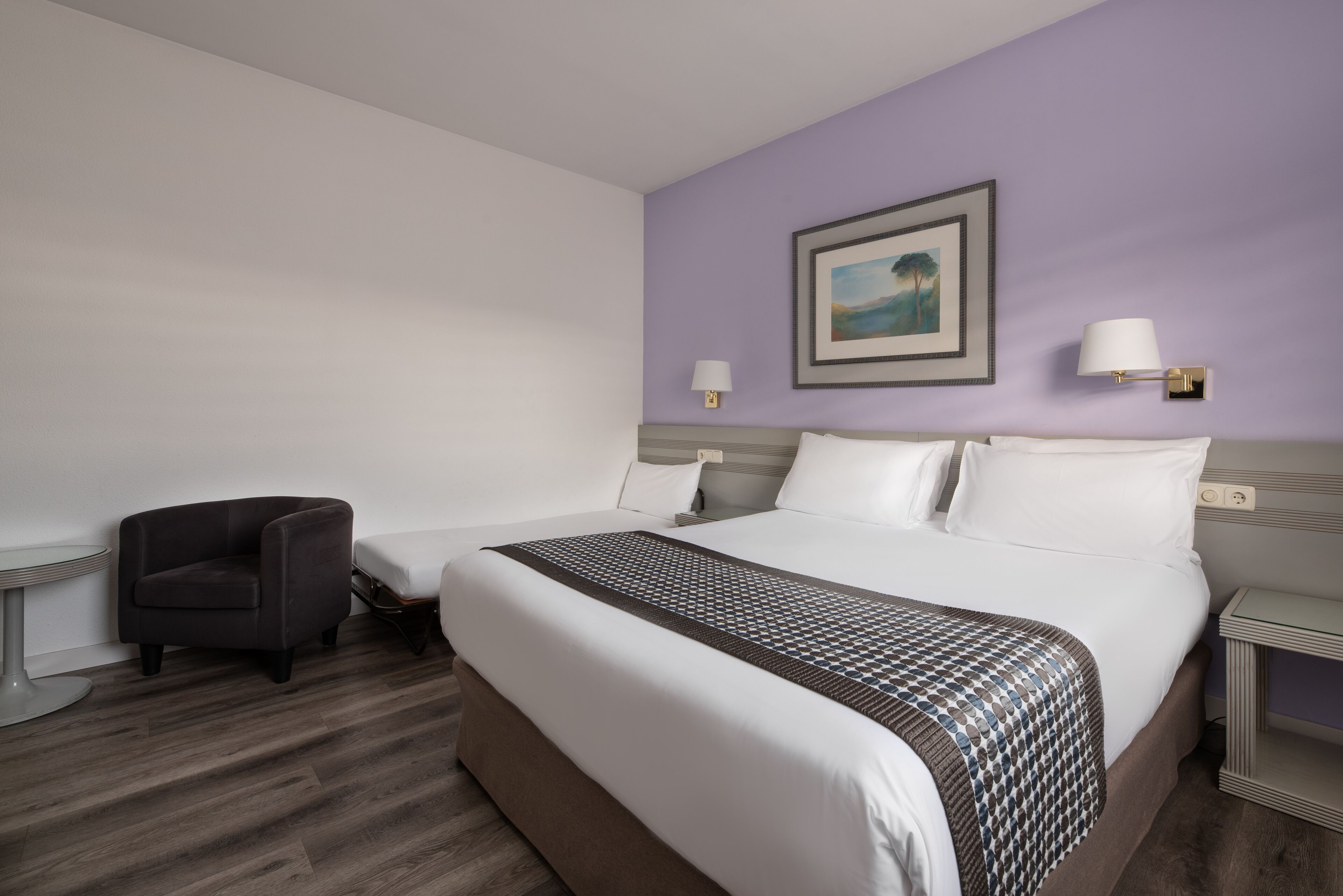 A hotel room with a large bed, a chair, a small table, and a wall-mounted lamp.
