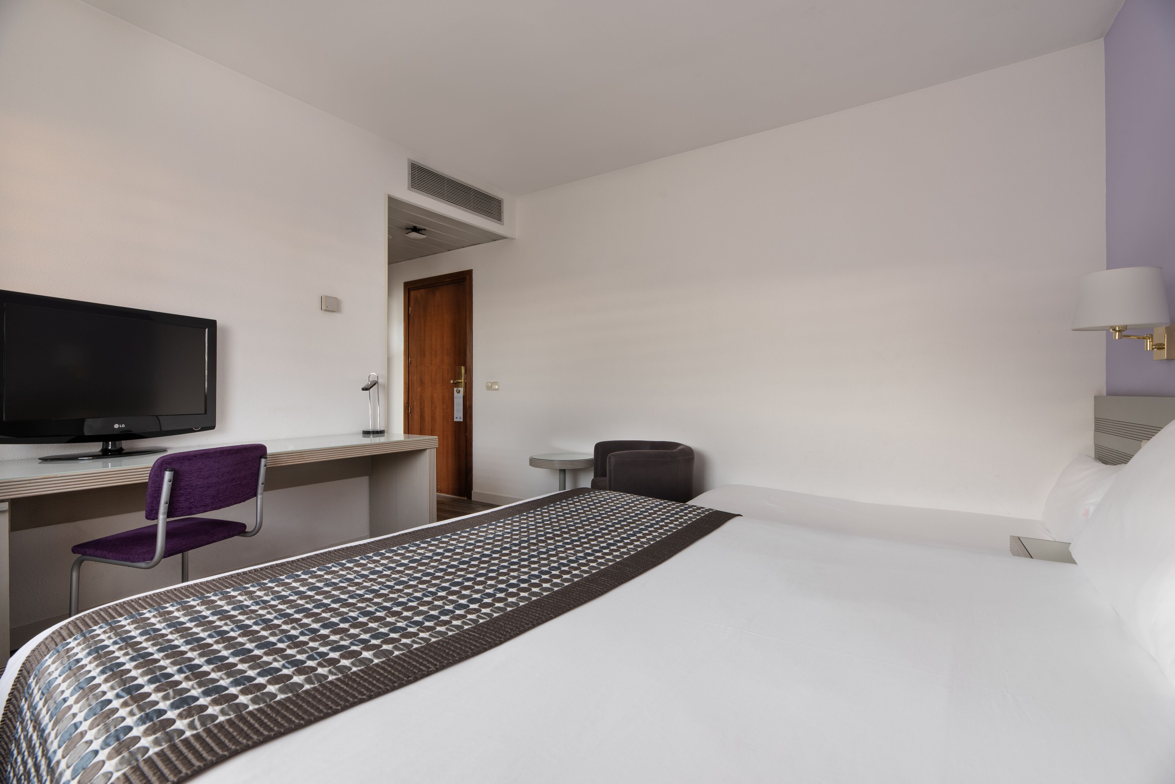 Una habitación de hotel con una cama, un escritorio con televisor, una silla y una mesita.