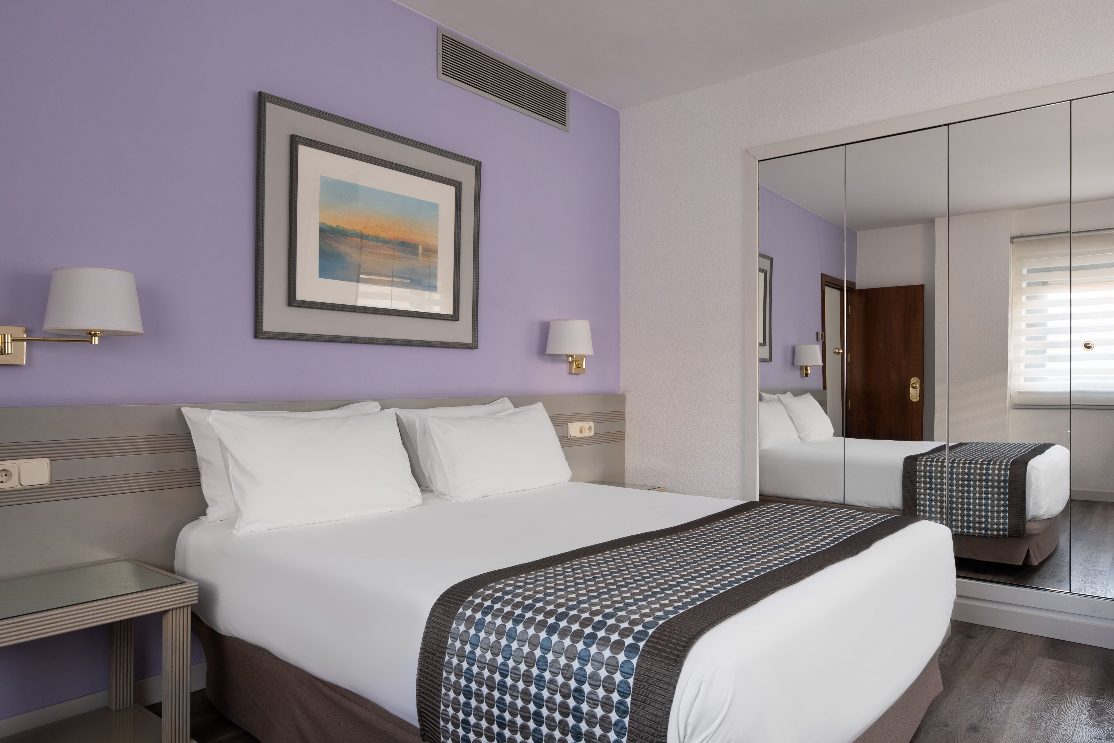 Habitación de hotel con una cama grande, dos mesitas de noche, un espejo y un cuadro en la pared.