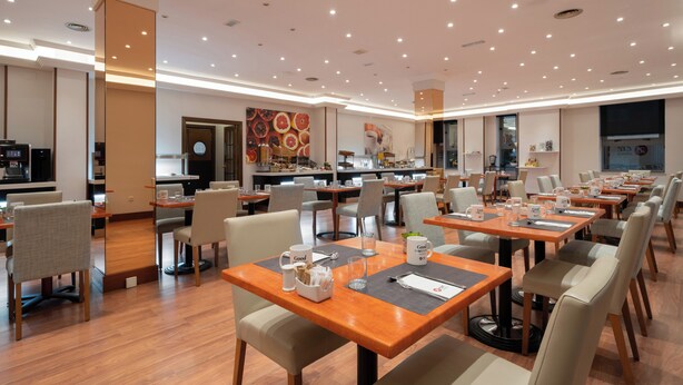 A modern hotel dining area with multiple tables and chairs, a kitchen in the background, and a large window. - Exe Salamanca (Salamanca)