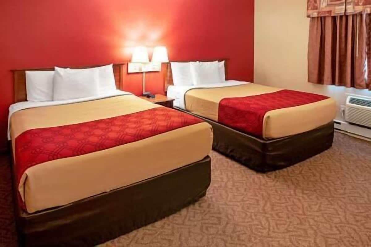 Two beds with red and beige bedding, a nightstand with a lamp, and a window with curtains.