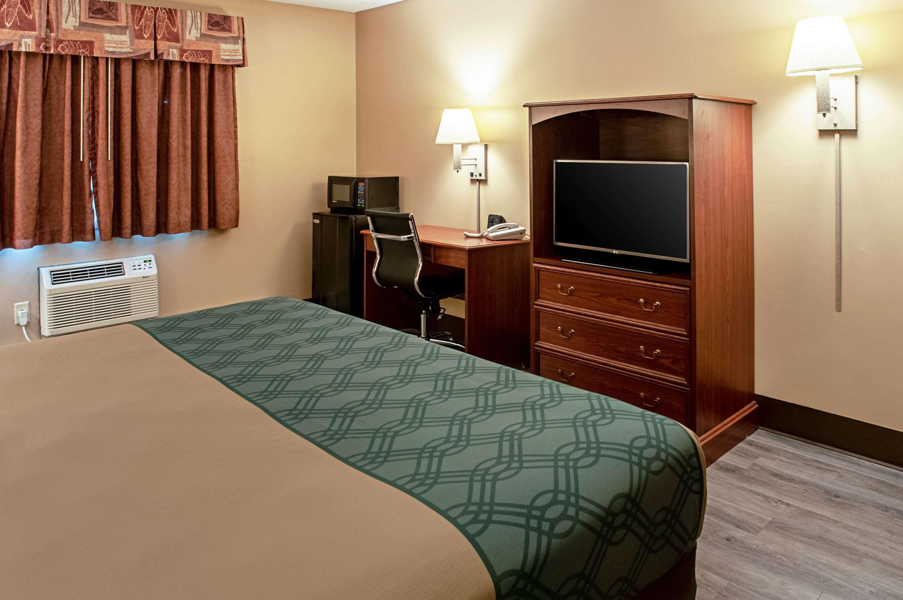 A hotel room with a bed, a desk with a computer, a television, a minibar, and a lamp.