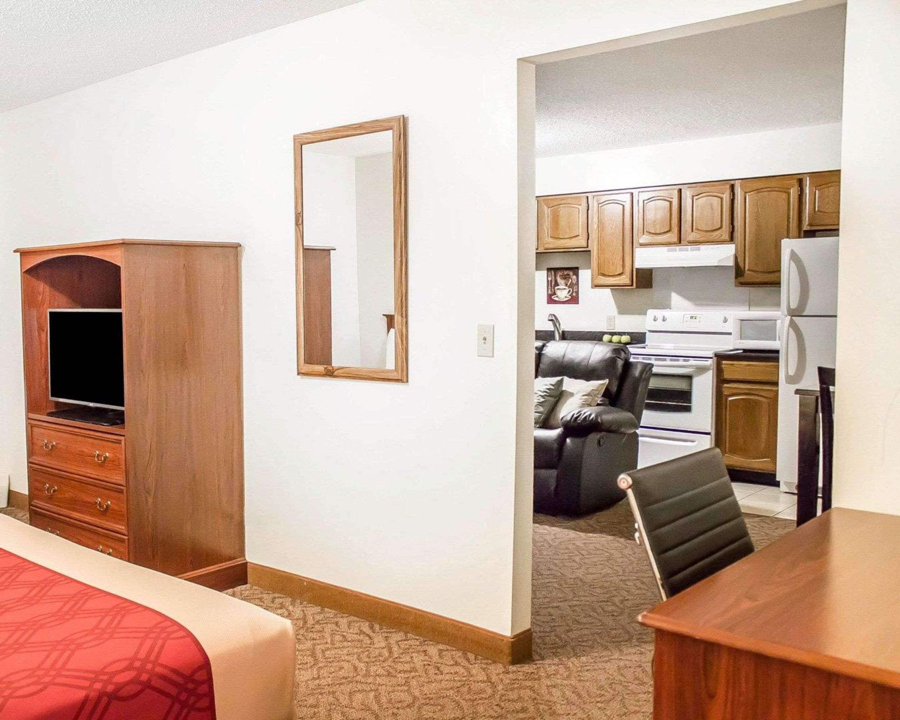 A hotel room with a wooden wardrobe, a television, a mirror, a bed with a red blanket, a small desk, and a kitchenette with a stove, microwave, and cabinets.