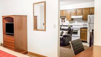 A hotel room with a wooden wardrobe, a television, a mirror, a bed with a red blanket, a small desk, and a kitchenette with a stove, microwave, and cabinets.
