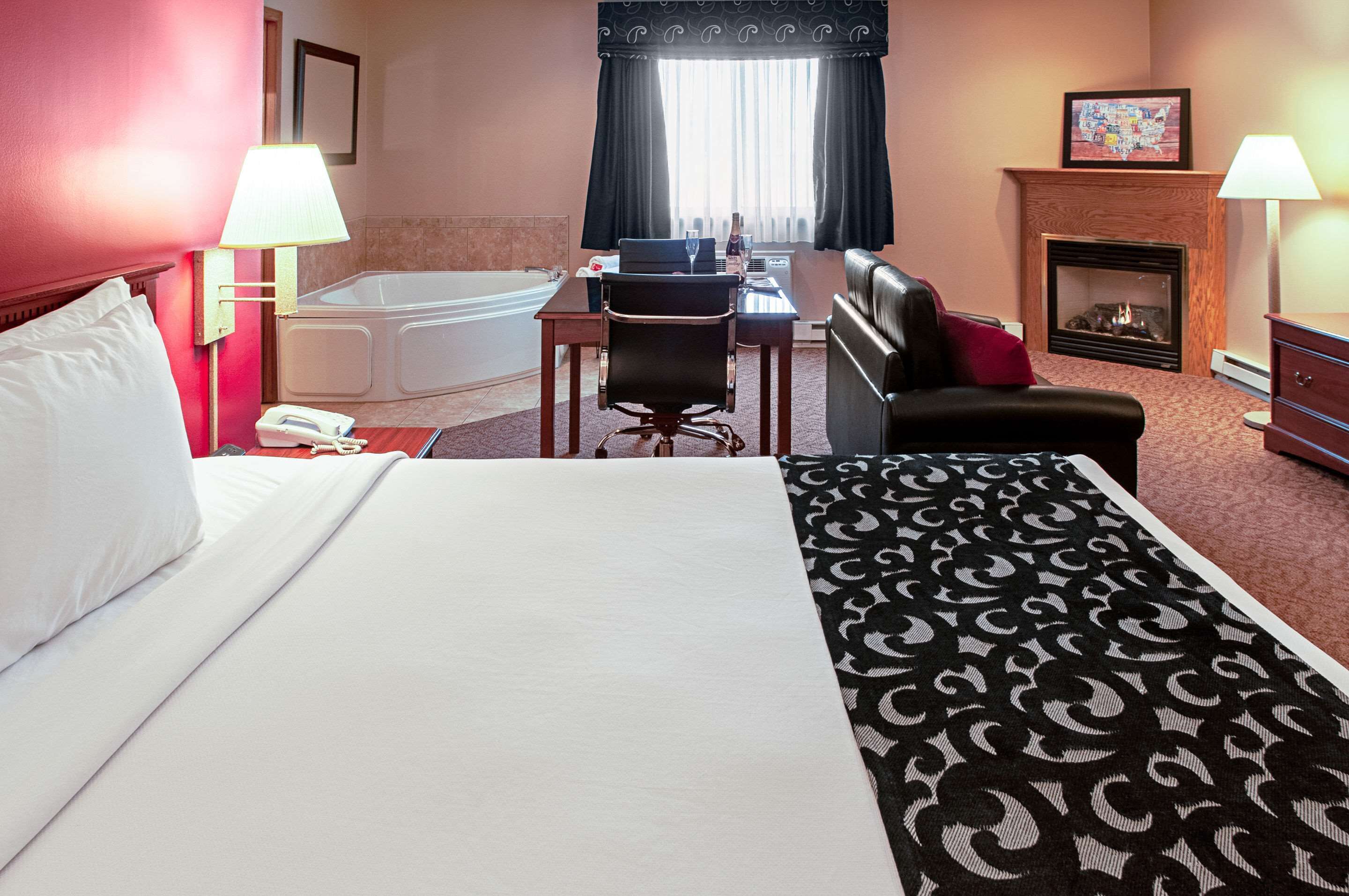 A hotel room with a large bed, a desk with a chair, a small table, a bathtub, a fireplace, and two lamps.