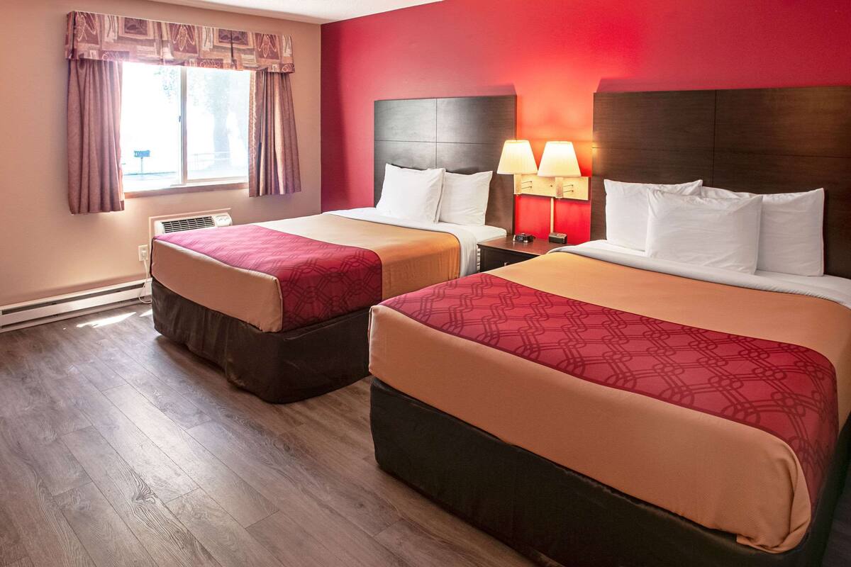 A hotel room with two beds, a window with curtains, a heater, and a nightstand with lamps.
