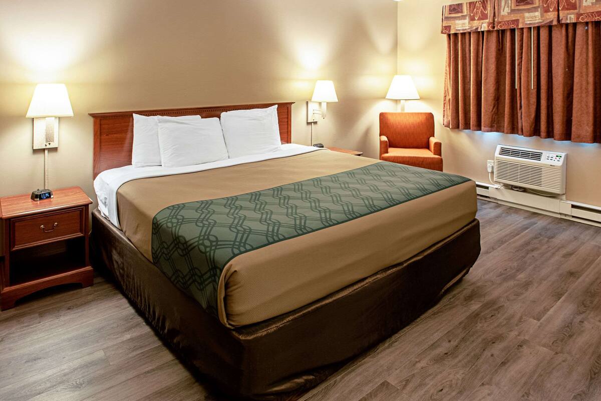 A hotel room with a large bed, two bedside lamps, a wooden nightstand, a chair, and an air conditioning unit.