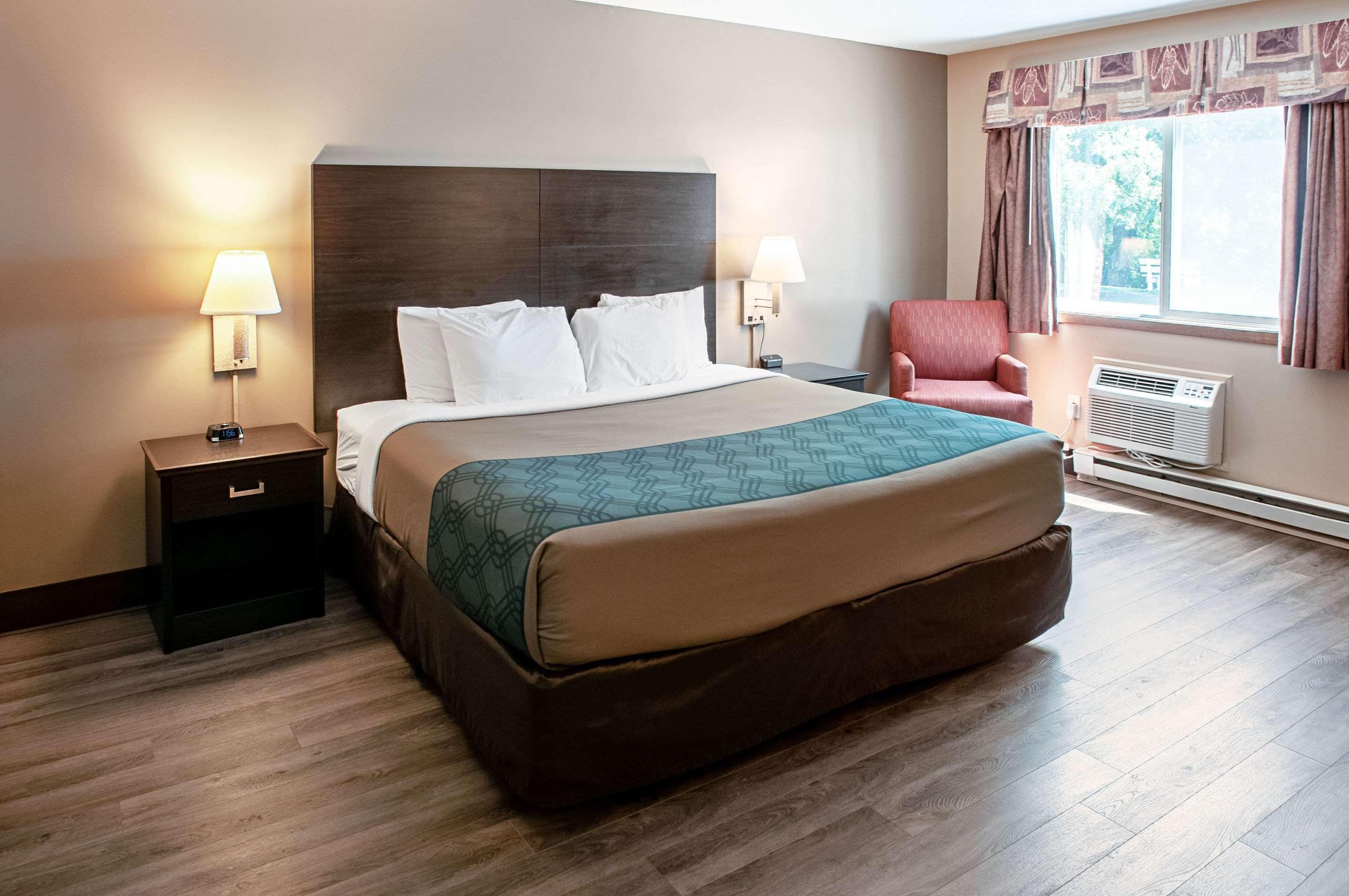 A hotel room with a large bed, two bedside tables with lamps, a red armchair, and a window with curtains.