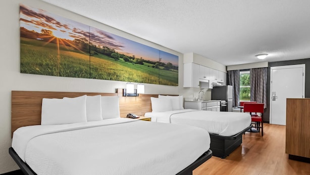 Une chambre d’hôtel avec deux lits, un grand tableau représentant un paysage, un bureau avec une chaise et un coin cuisine.