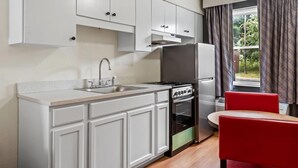 Une cuisine compacte avec des meubles blancs, un évier en acier inoxydable, une cuisinière et un réfrigérateur. Il y a une fenêtre avec des rideaux et une chaise rouge dans le coin repas.