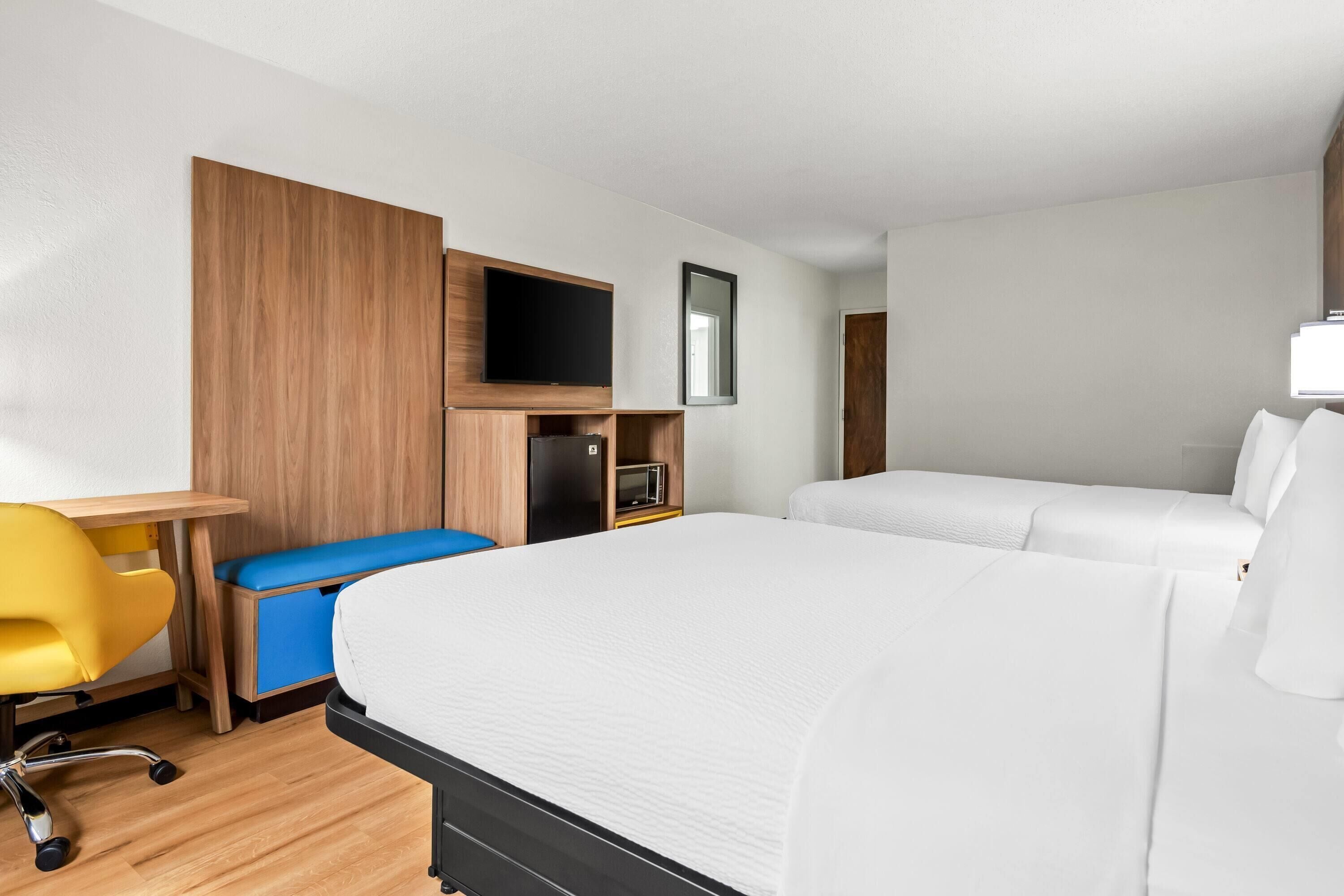 A hotel room with a large bed, a desk with a chair, a TV, and a small seating area with cushions.