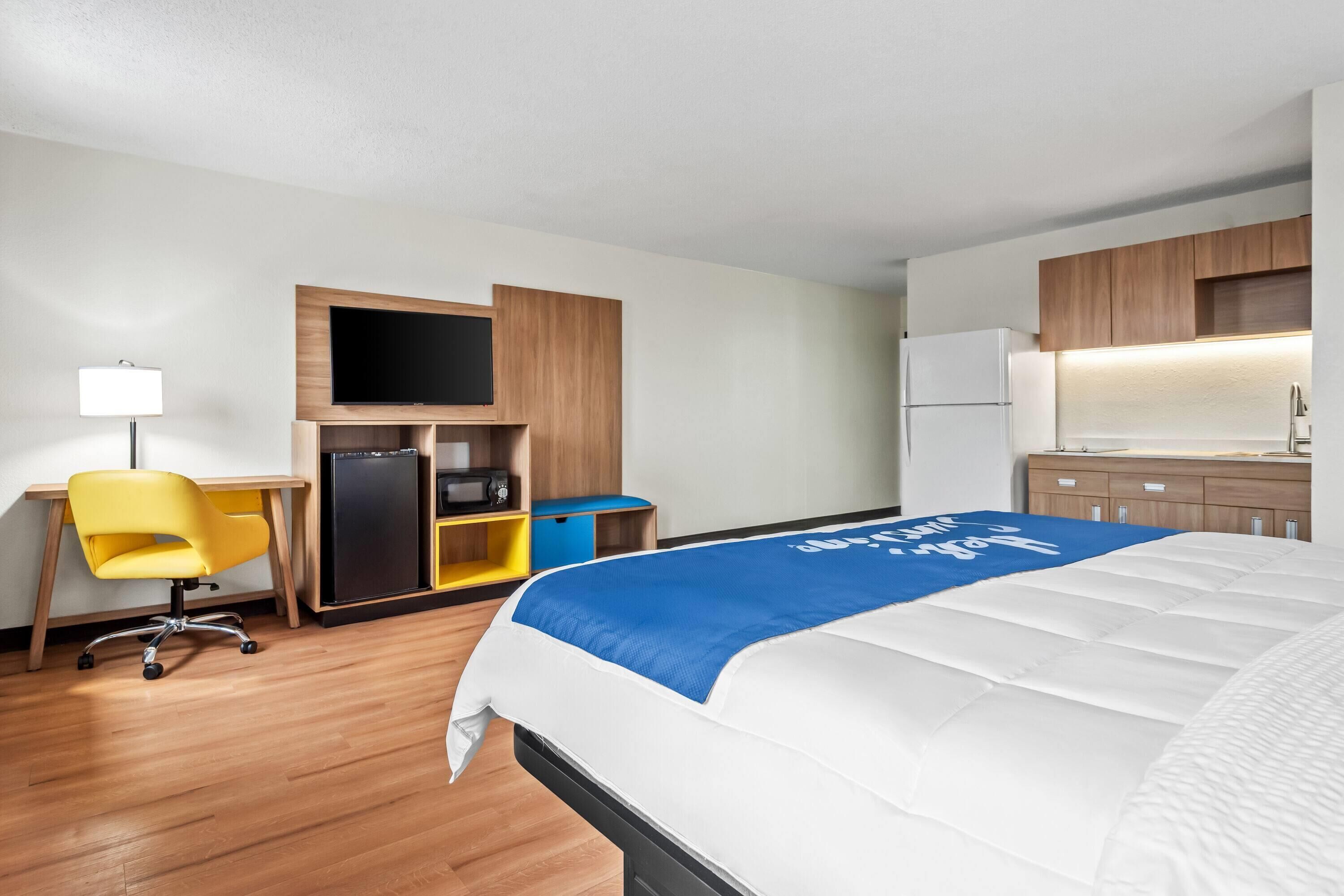 A hotel room with a bed, a desk, a chair, a TV, a microwave, a mini-fridge, and a kitchenette.