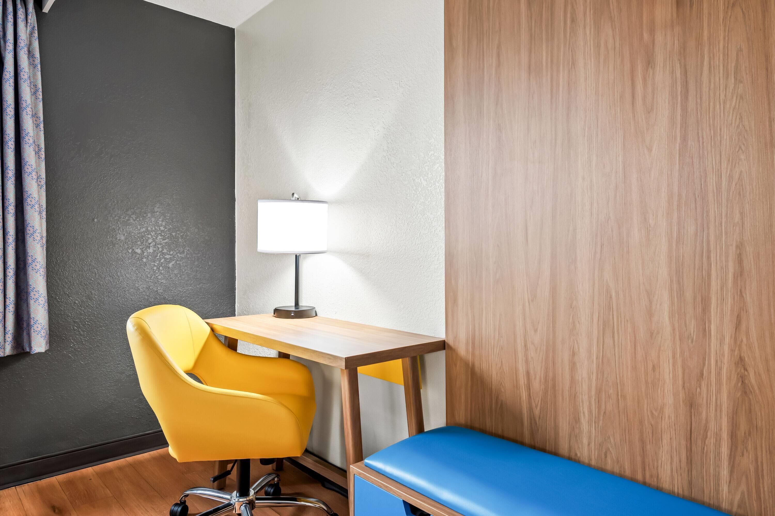 Um quarto com uma escrivaninha de madeira, uma cadeira amarela, um banco azul e uma luminária.