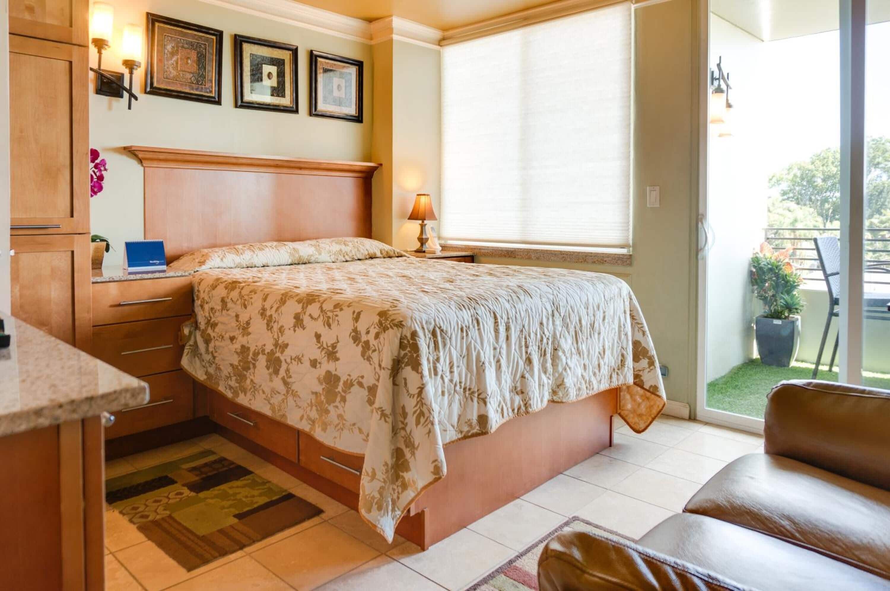 A bedroom with a bed, a nightstand, a lamp, a window with blinds, and a view of the outdoors.