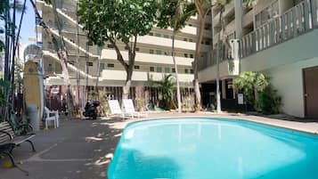 A swimming pool surrounded by a multi-story building with balconies, trees, and a few lounge chairs.