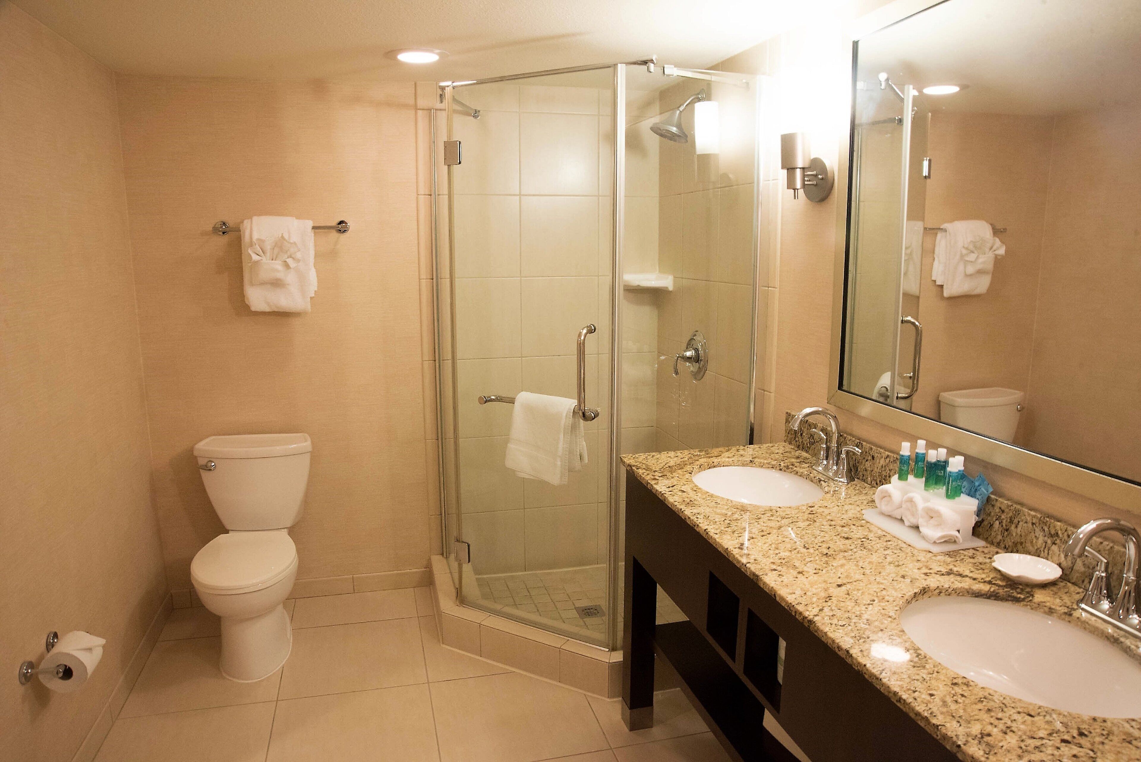 A hotel bathroom with a glass-enclosed shower, a toilet, and a vanity with two sinks and a mirror.