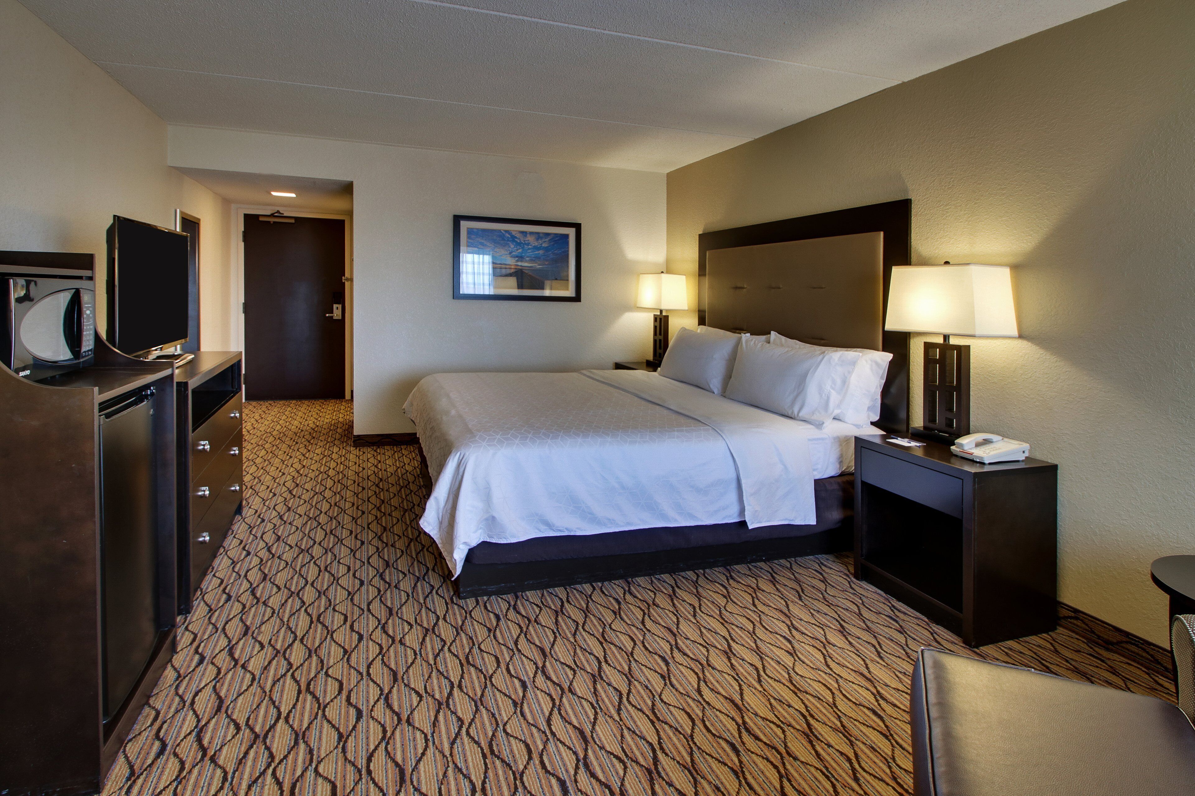 A hotel room with a bed, a desk with a lamp, a television, and a patterned carpet.