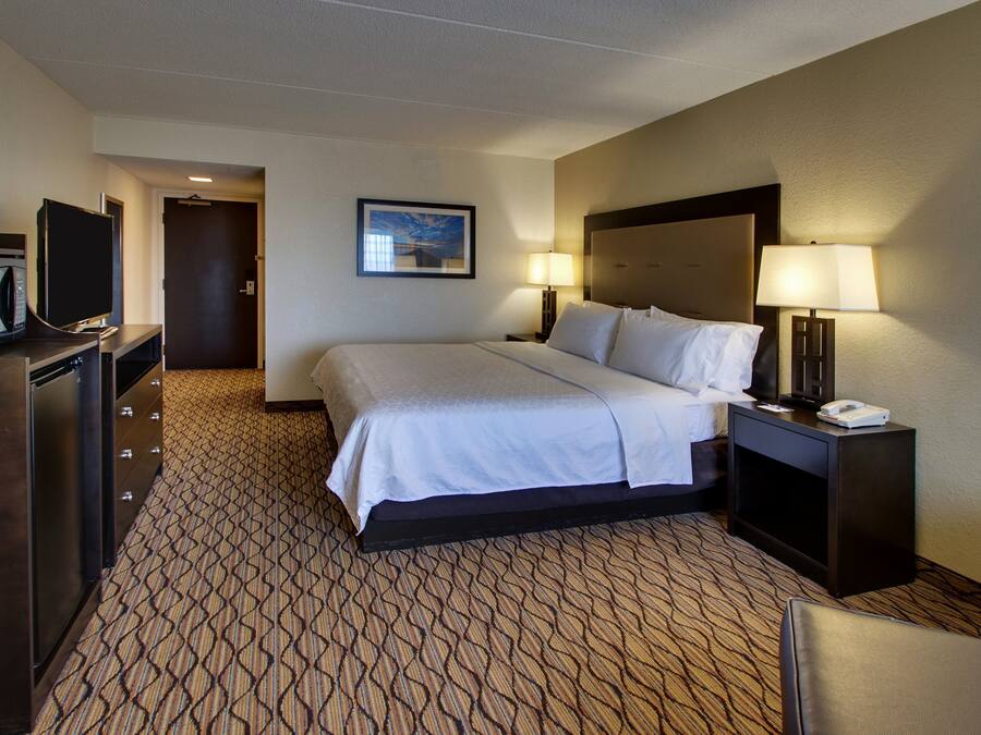 A hotel room with a bed, a desk with a lamp, a television, and a patterned carpet.