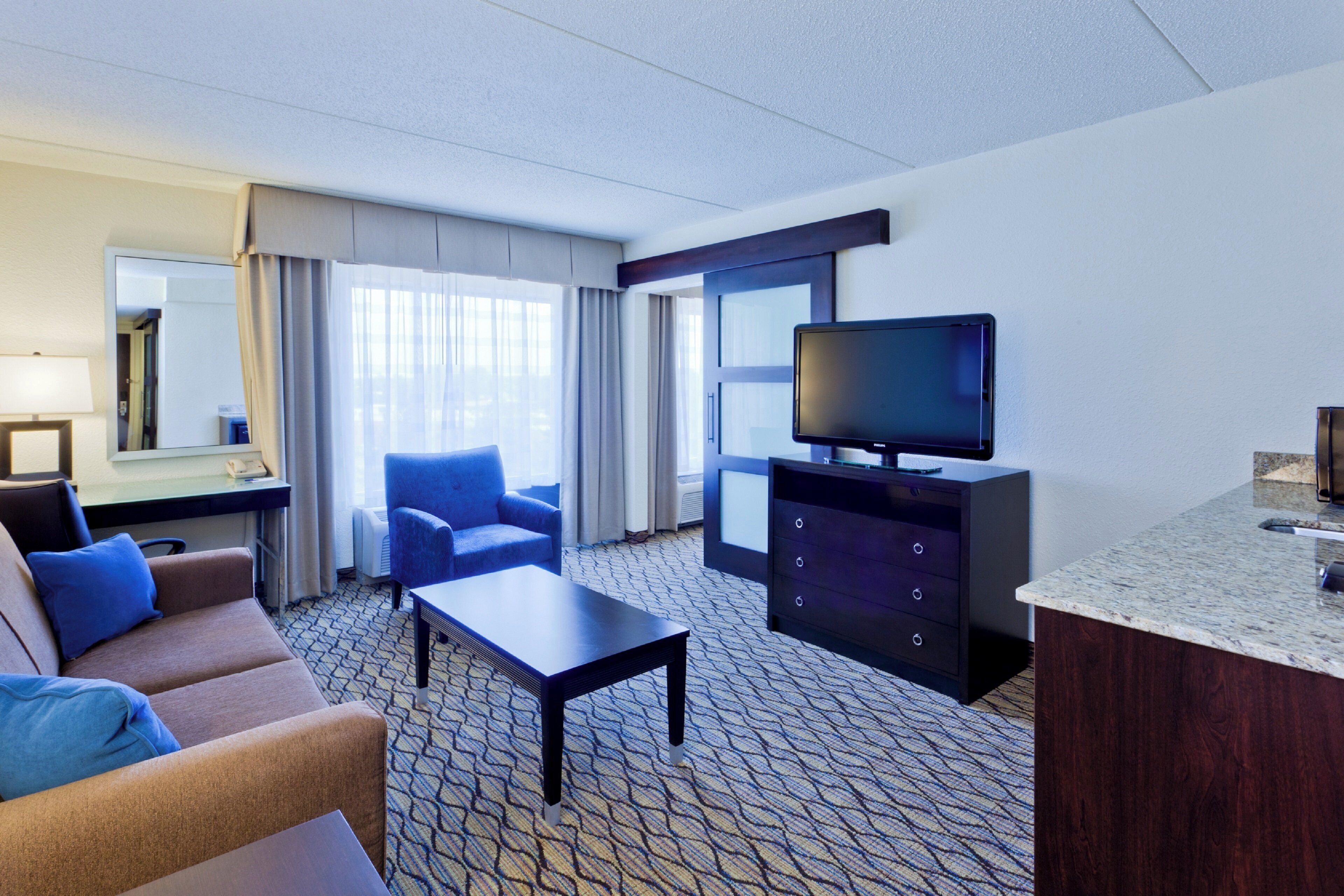 A hotel room with a sofa, armchair, TV, dresser, and a desk with a lamp.