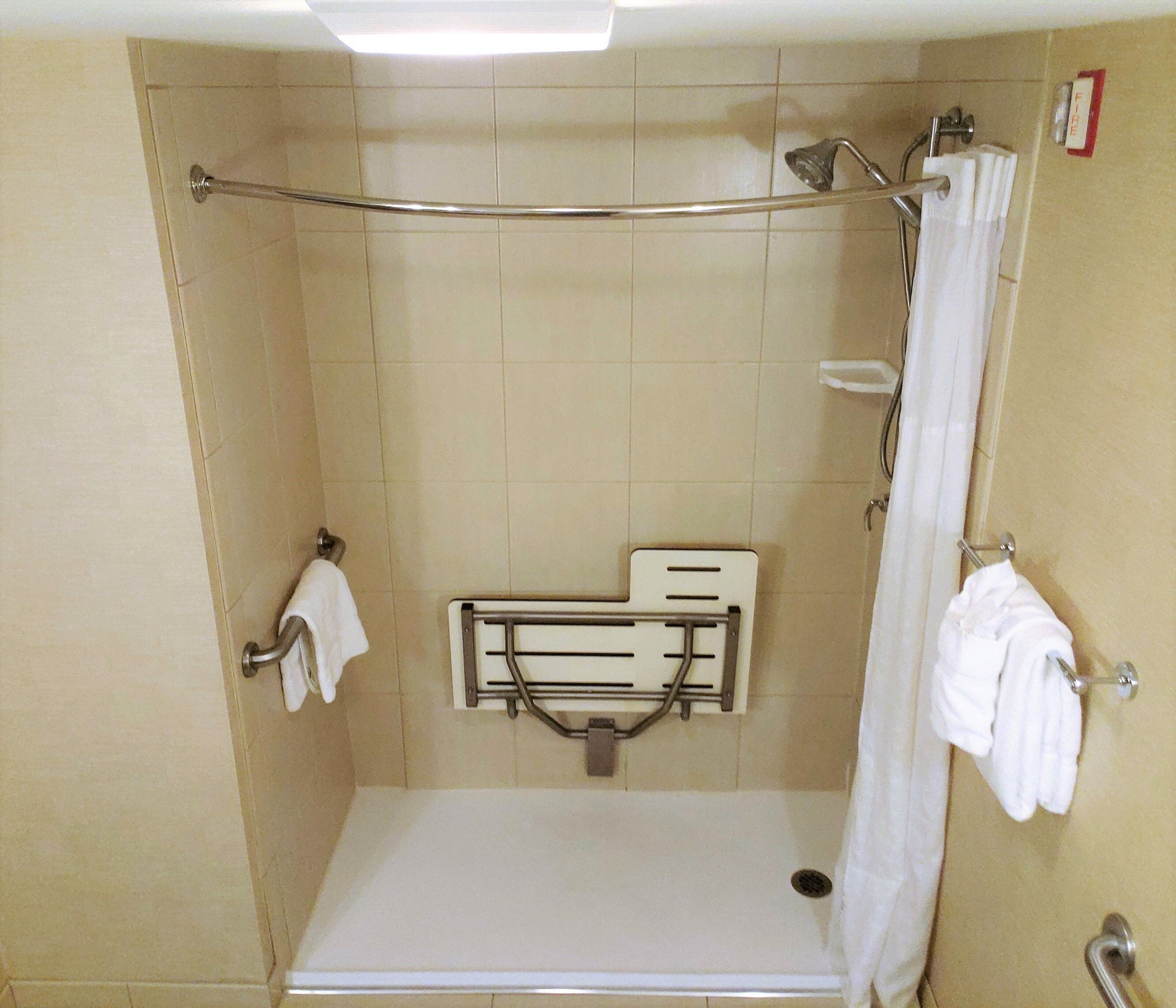 Un box de ducha pequeño y cerrado con un asidero, un toallero blanco y un detector de incendios.