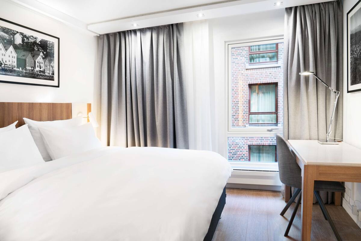 A hotel room with a bed, a desk, a chair, a window with curtains, and a framed picture on the wall.