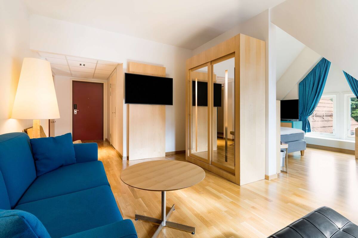 A modern hotel room with a blue sofa, a round wooden table, a TV mounted on the wall, and a large mirror.