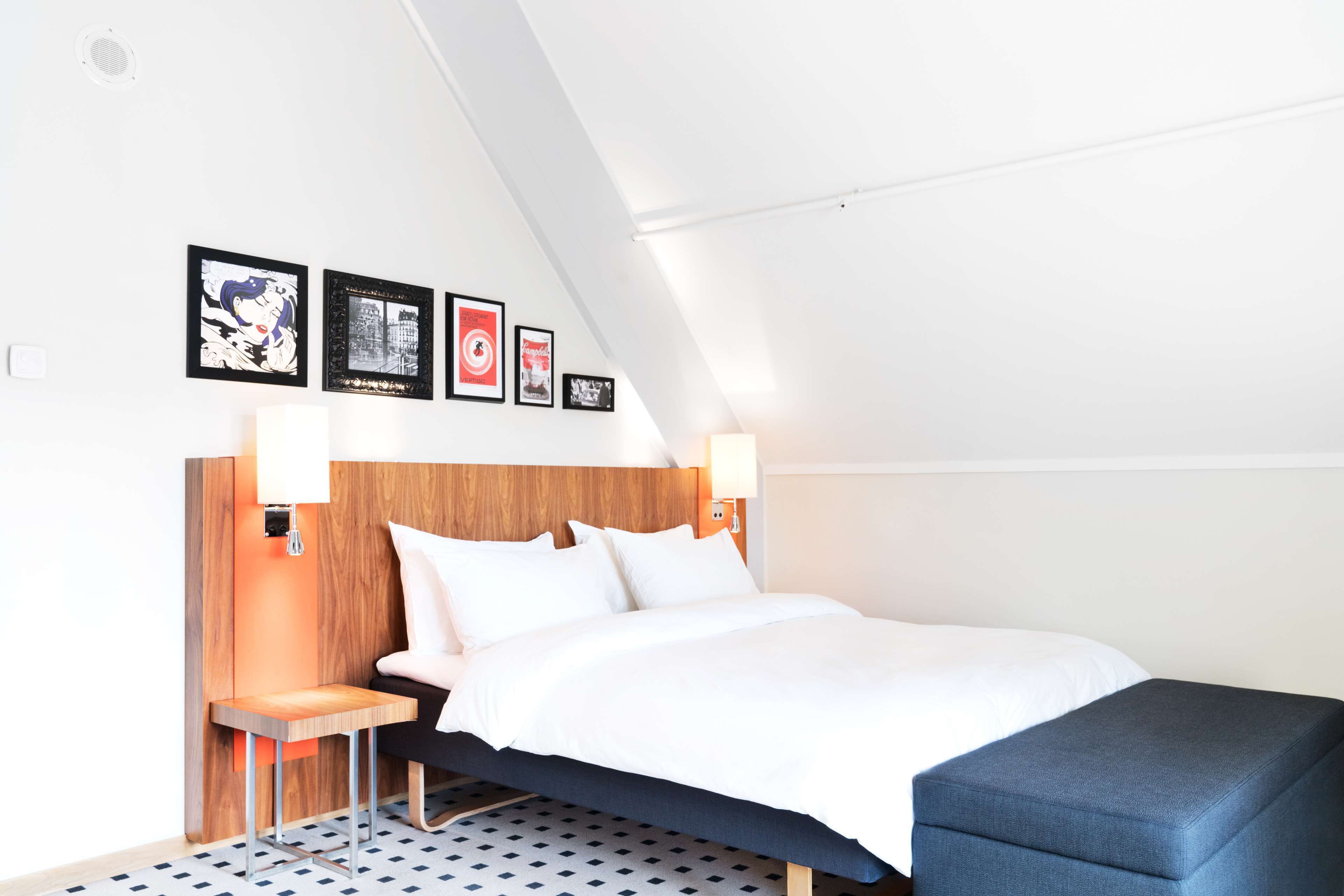 Una habitación de hotel moderna con cabecera de madera, cama con ropa blanca, una mesita con lámpara y un banco.