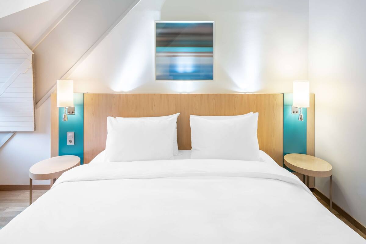 A modern hotel room with a large bed, two bedside tables, a headboard, and wall-mounted lamps.