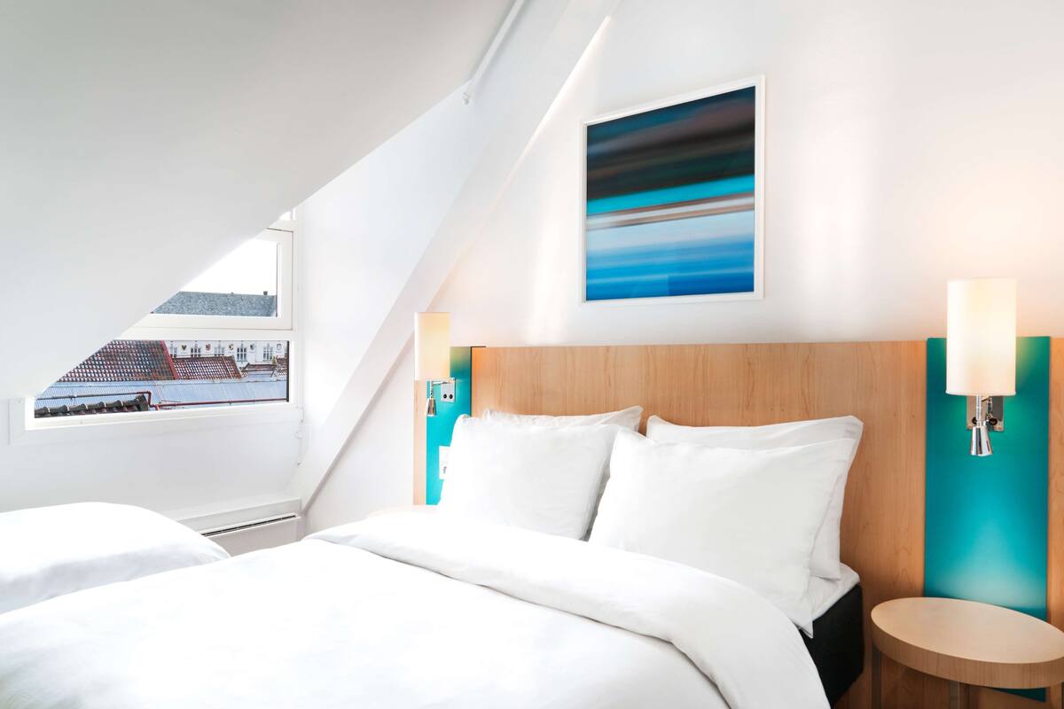 A modern hotel room with a large bed, a wooden headboard, a view of the city through a skylight, and a painting on the wall.