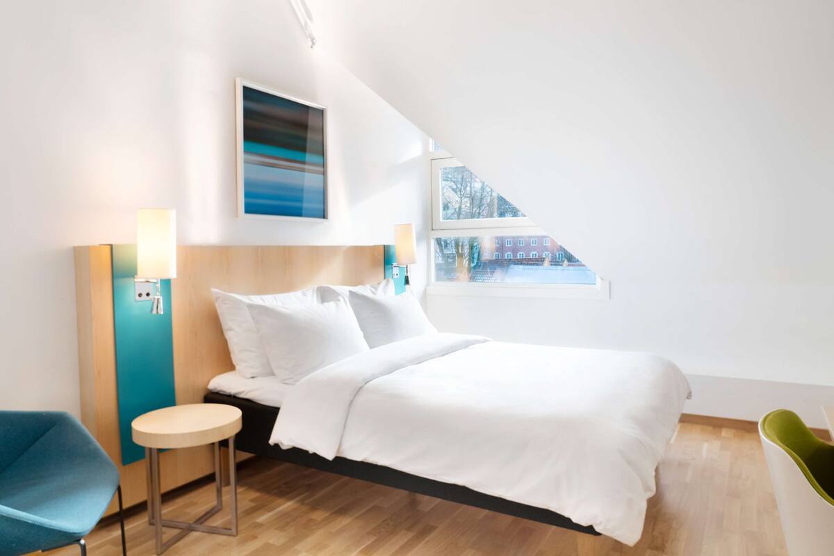 A modern hotel room with a bed, a small round table, a blue chair, and a window with a view of buildings.