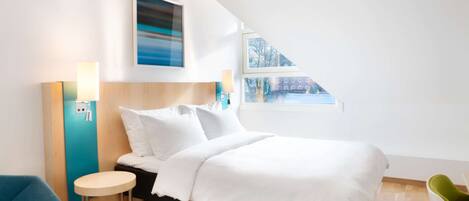 A modern hotel room with a bed, a small round table, a blue chair, and a window with a view of buildings.