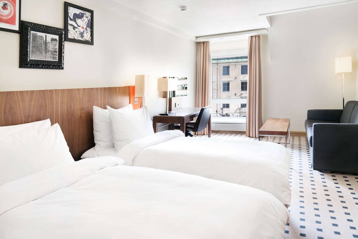 A hotel room with two beds, a desk, a chair, a sofa, and a view of the city through the window.
