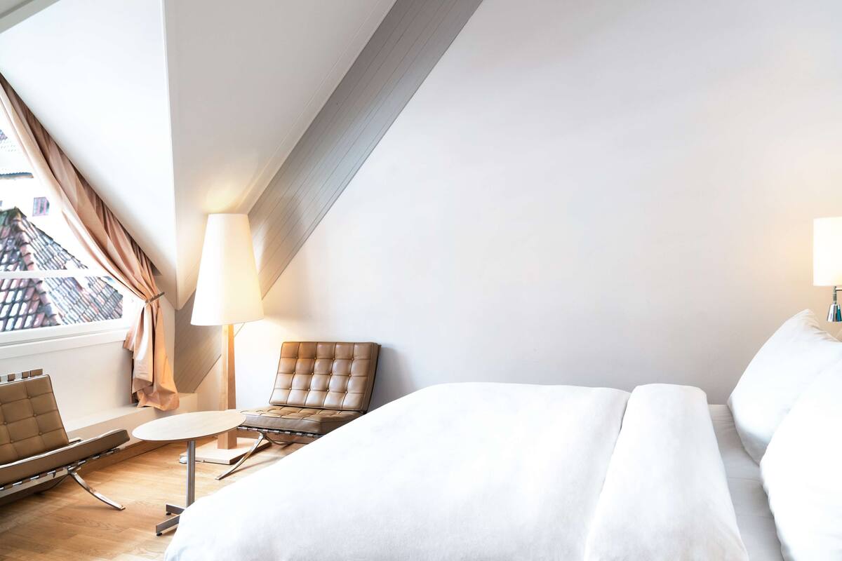 A modern hotel room with a bed, a chair, a small table, and a lamp.