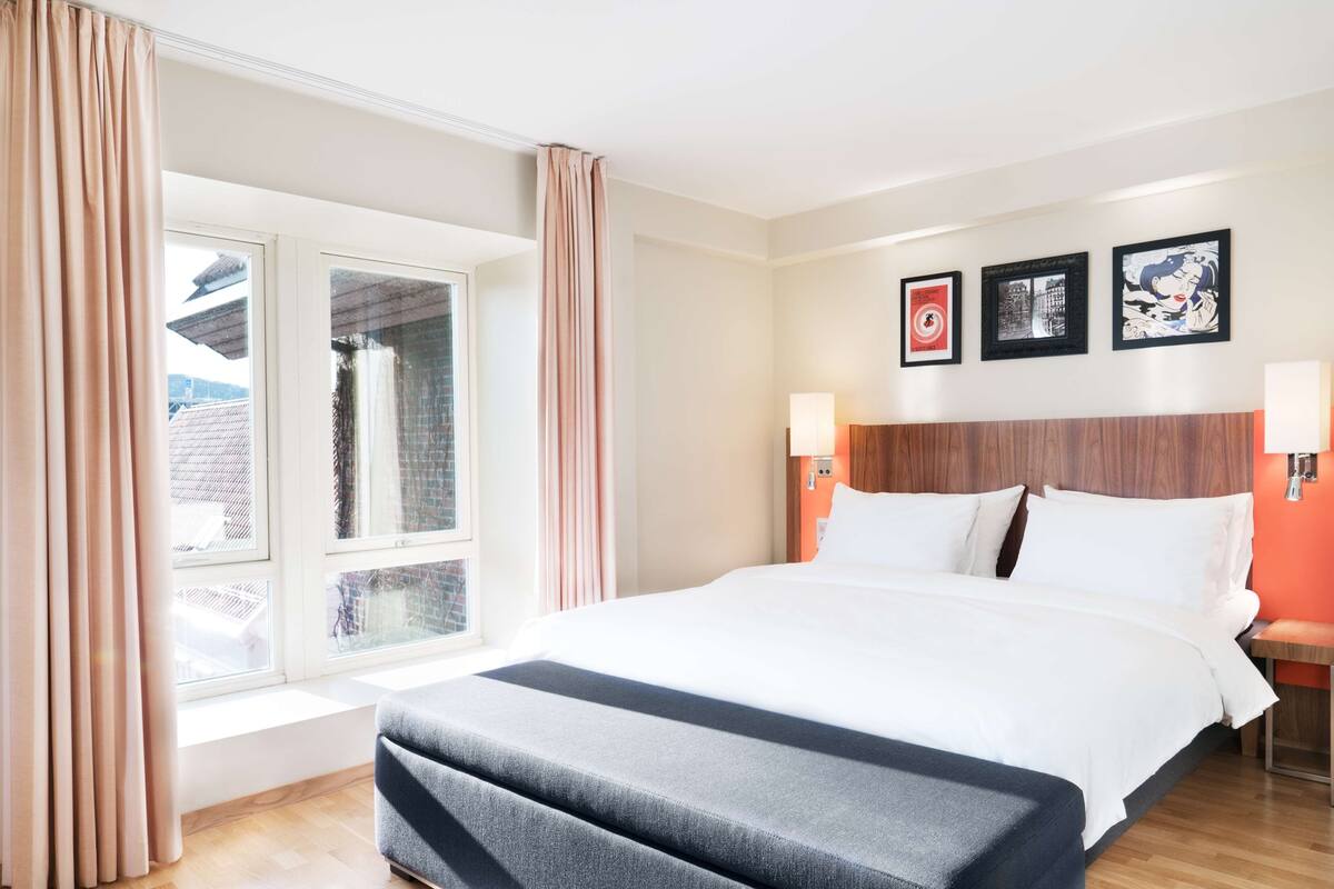 A hotel room with a large bed, a window with curtains, a wooden headboard, and wall-mounted lamps.