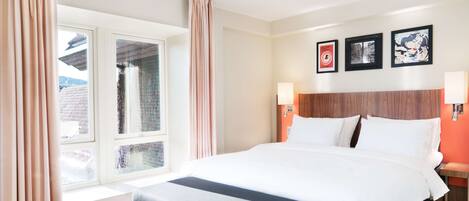 Habitación de hotel con una cama grande, una ventana con cortinas, un cabecero de madera y tres cuadros enmarcados en la pared.
