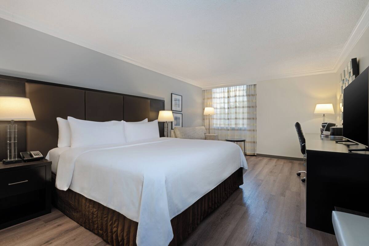 Ein Hotelzimmer mit einem großen Bett, einem Schreibtisch und einem Stuhl. Zwei Nachttische mit Lampen und ein Telefon sind vorhanden. Der Raum verfügt über Holzboden und ein Fenster mit Vorhängen.