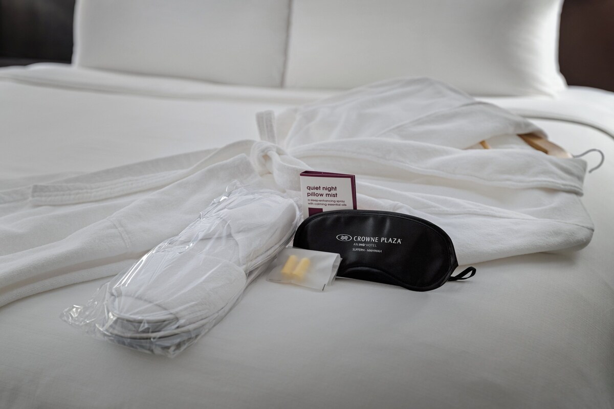 A neatly made bed with white linens, a pillow mist spray bottle, and a black eye mask.