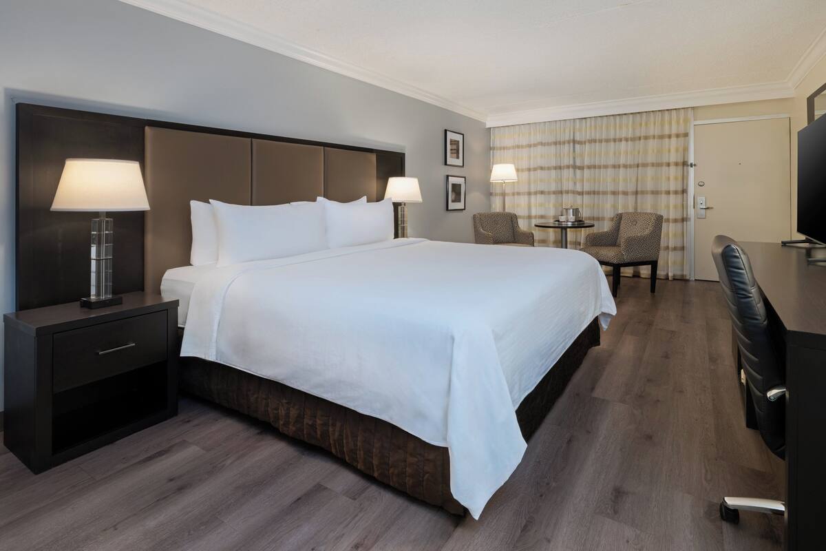 Ein Hotelzimmer mit einem großen Bett, einem Nachttisch mit Lampe, einem Schreibtisch mit Stuhl und einem Fernseher.
