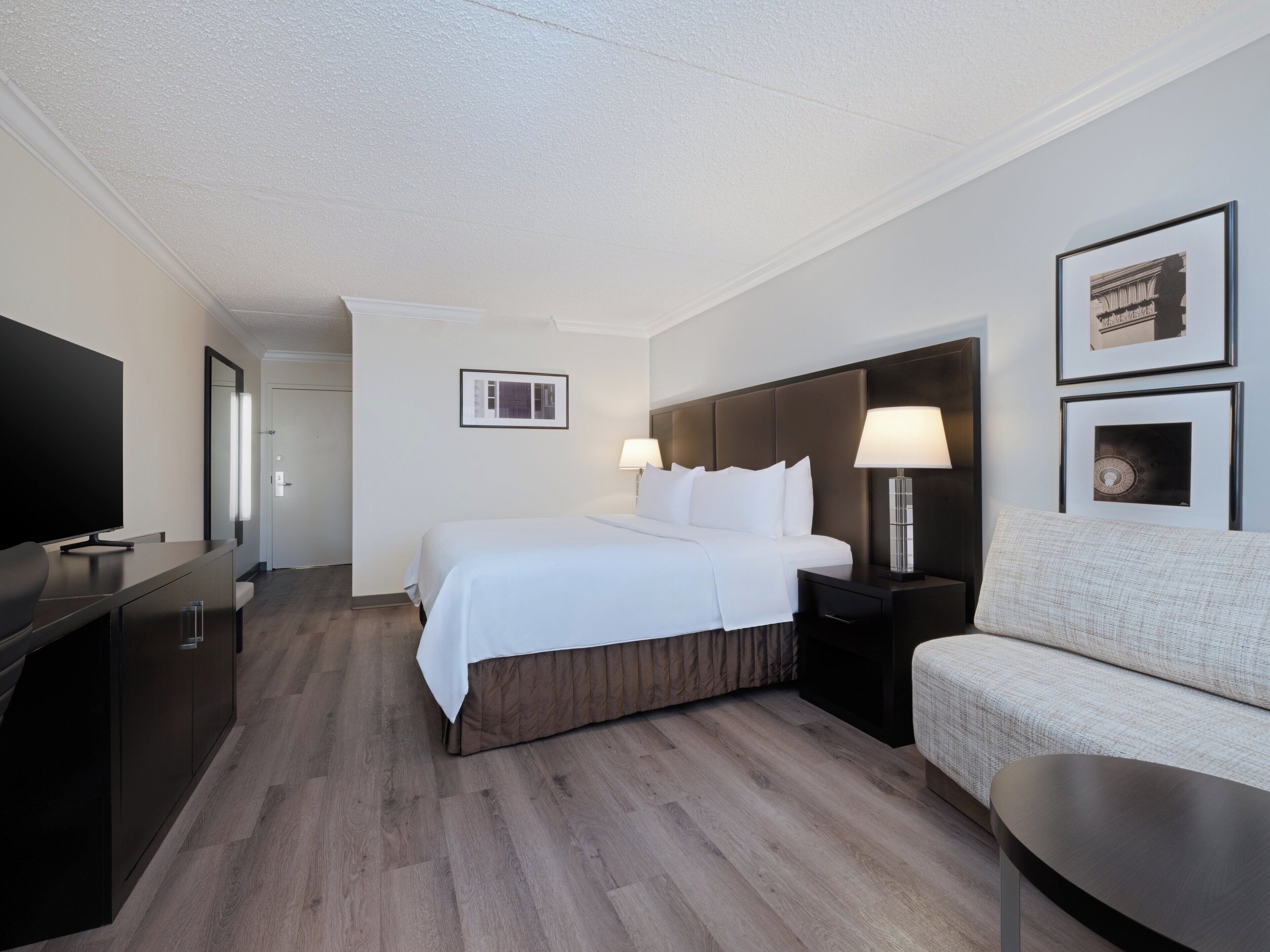 Une chambre d’hôtel avec un lit, des tables de chevet, une télévision, un canapé et des tableaux encadrés au mur.