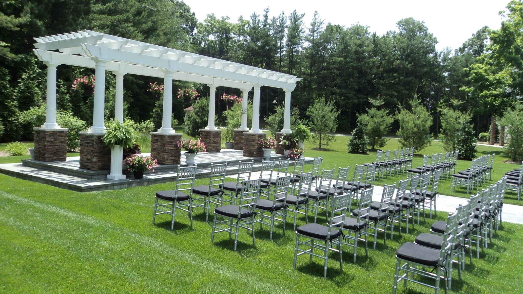 A white pergola with columns and stone bases, surrounded by chairs and potted plants.
