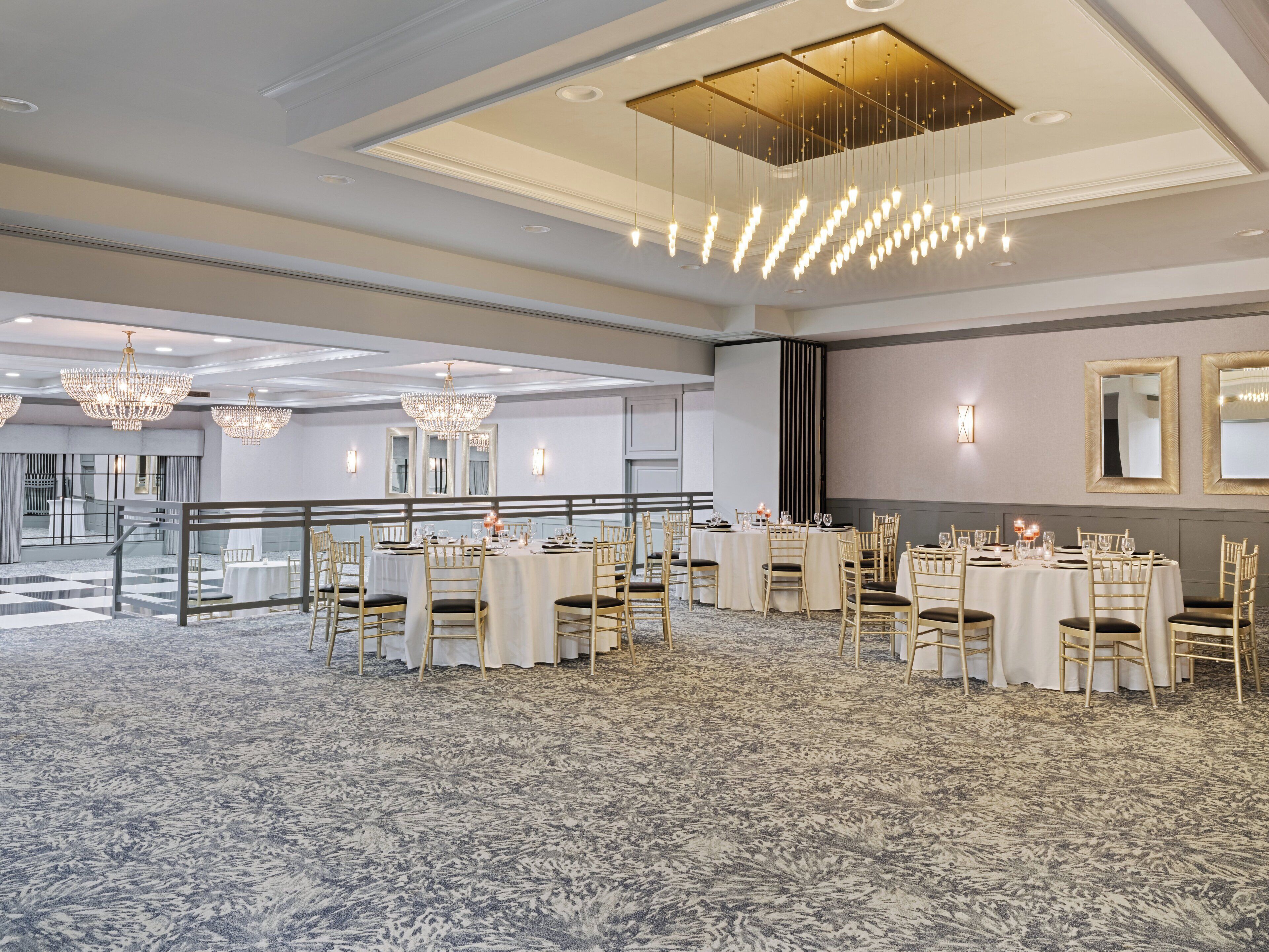 A spacious banquet hall with multiple round tables, elegant chandeliers, and a patterned carpet.