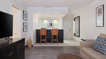 Un salon moderne avec un téléviseur à écran plat, un meuble en bois foncé, un canapé beige agrémenté d’un coussin à motifs et une petite table basse. On y trouve deux chaises en bois près d’un comptoir de cuisine, avec un pare-feu blanc et des luminaires suspendus.