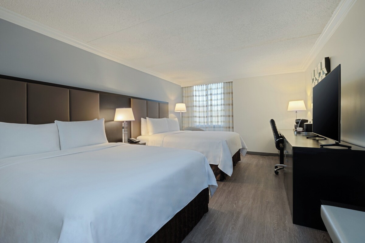 A hotel room with two beds, a desk, a TV, and a window with curtains.