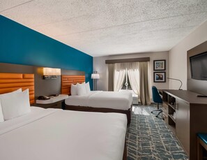 Ein Hotelzimmer mit zwei Betten, einem Schreibtisch mit Lampe, einem Fernseher und einem Fenster mit Vorhängen.