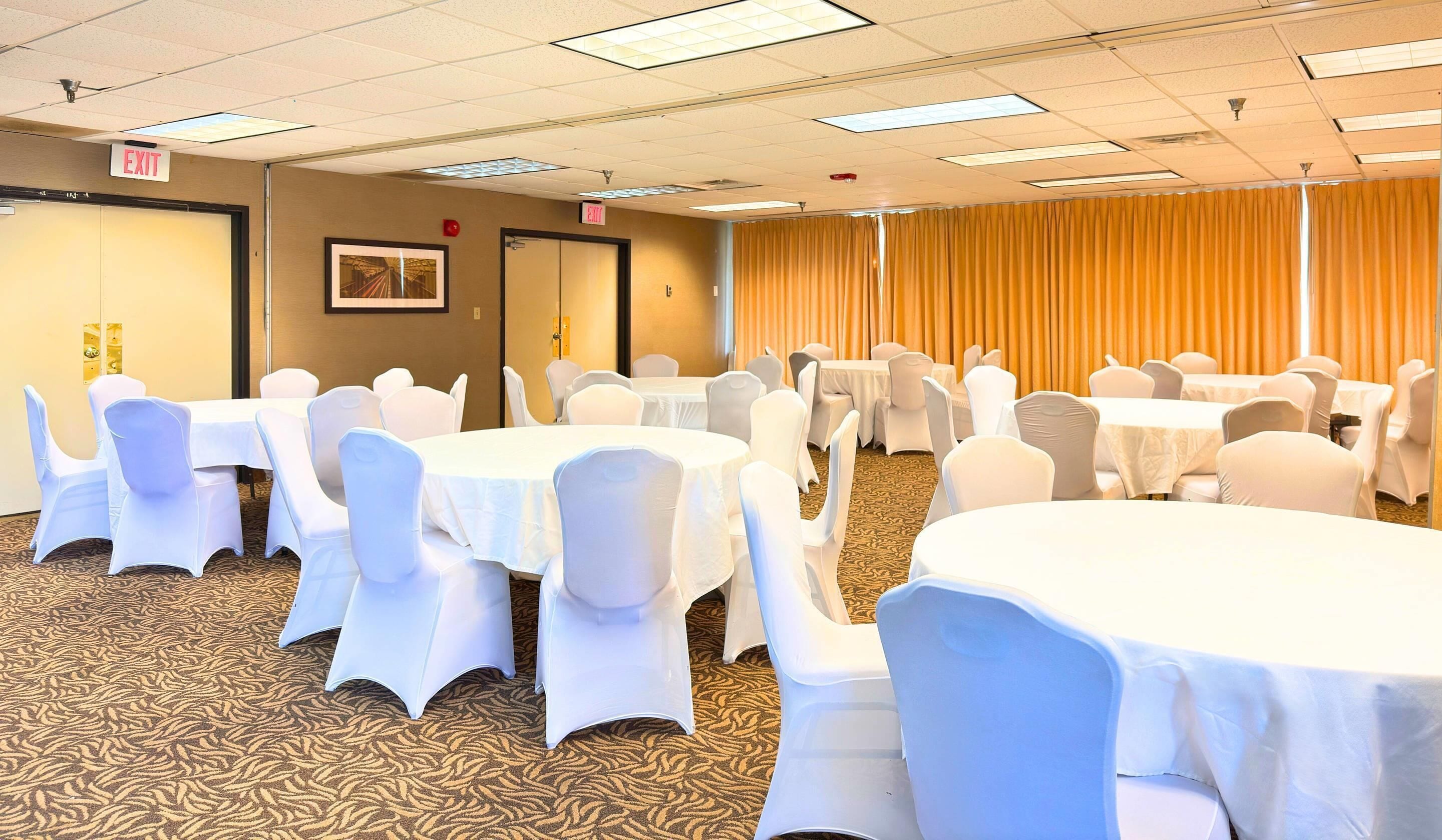 A conference room with multiple round tables and chairs, carpeted floor, exit signs, and a curtain backdrop.
