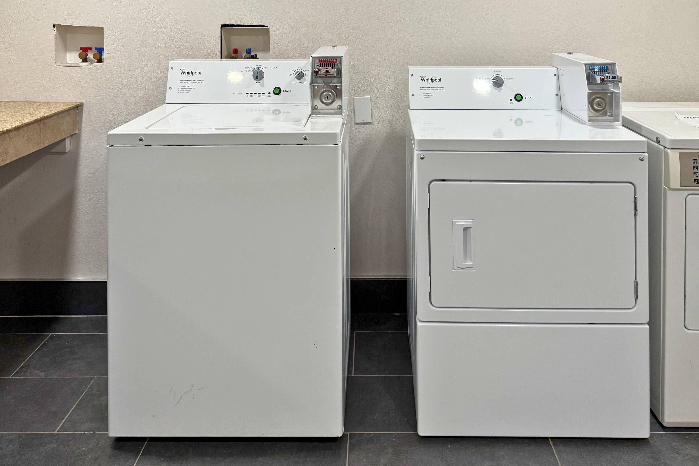 Dos lavadoras Whirlpool de carga frontal en un lavadero con una encimera y un estante pequeño con botellas.