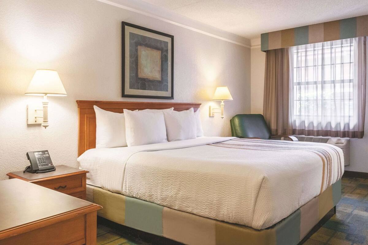 A hotel room with a bed, bedside lamps, a telephone, a chair, and a painting on the wall.